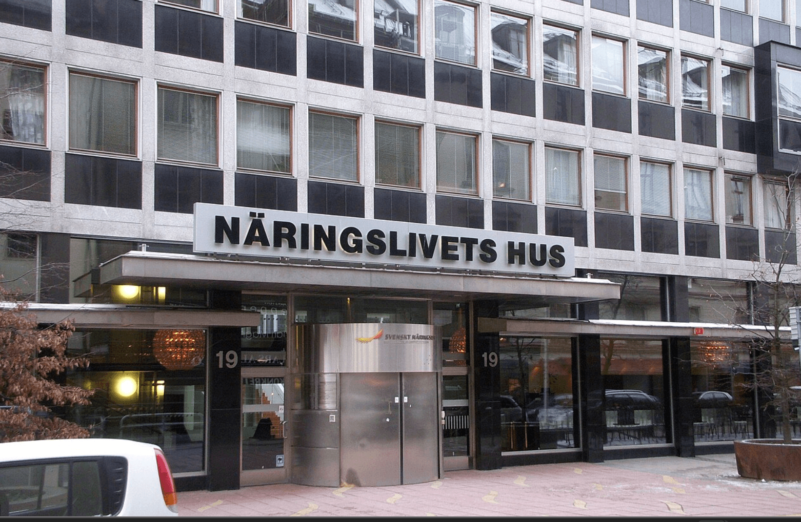 Bilden visar entrén till Näringslivets Hus med en modern fasad och stora fönster. Entrén har nummer 19 och en bil parkerad framför byggnaden.