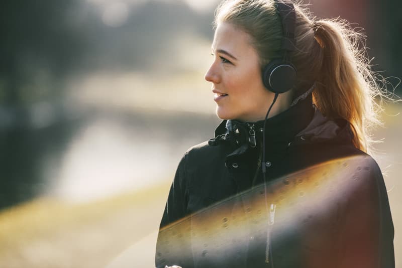 Kvinna i jacka och hörlurar ser åt sidan under en promenad utomhus, med mjukt motljus och ljusreflexer i bilden.
