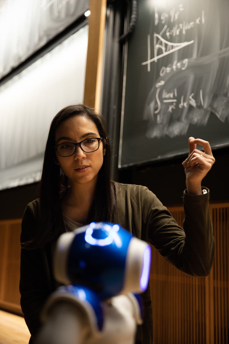 Kvinna med glasögon står vid en griffeltavla med matematiska formler och gestikulerar mot en liten blå robot i förgrunden.