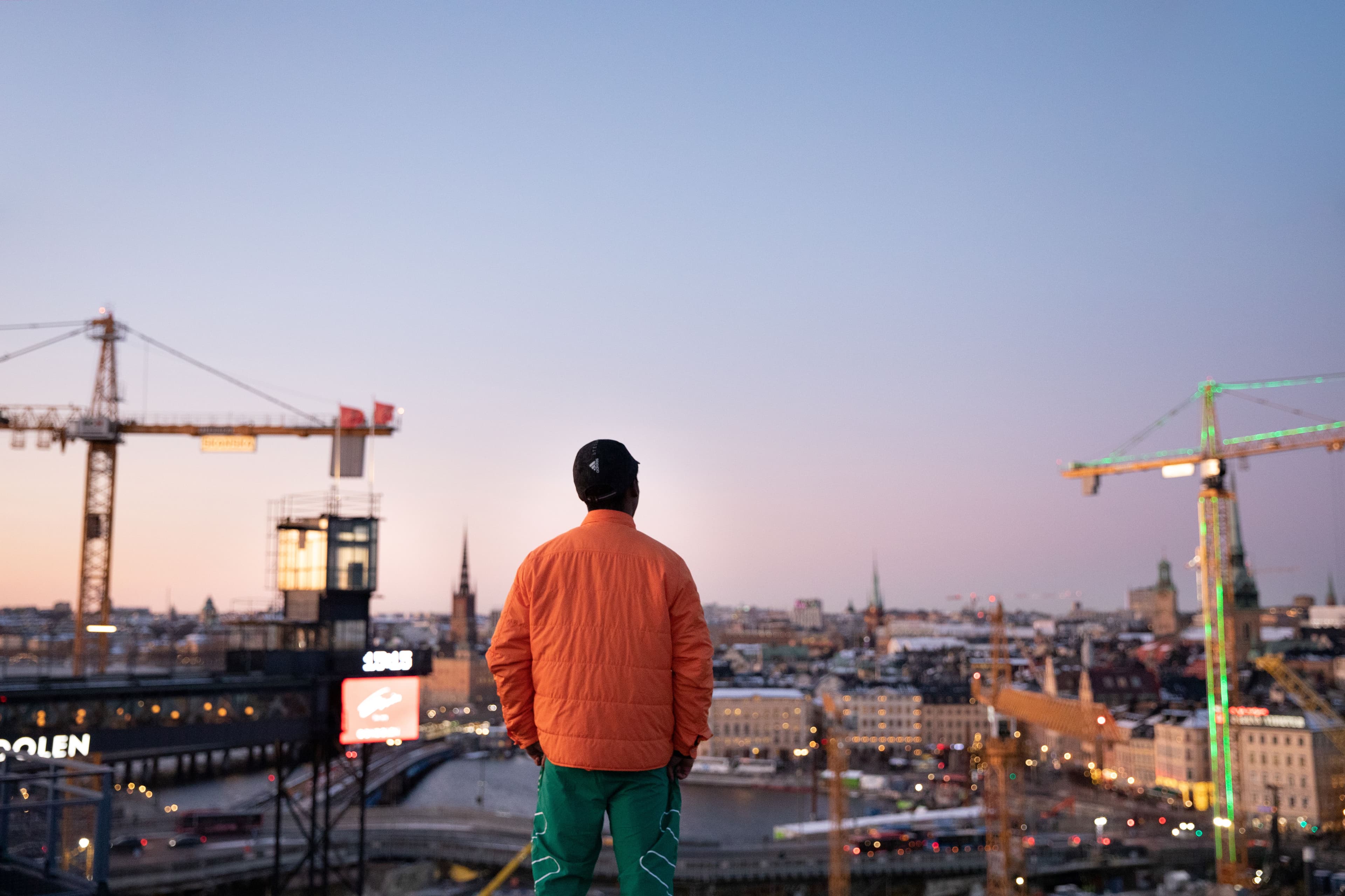 Person i orange jacka står med ryggen mot kameran och blickar ut över Stockholms skyline med byggkranar i skymningsljus.